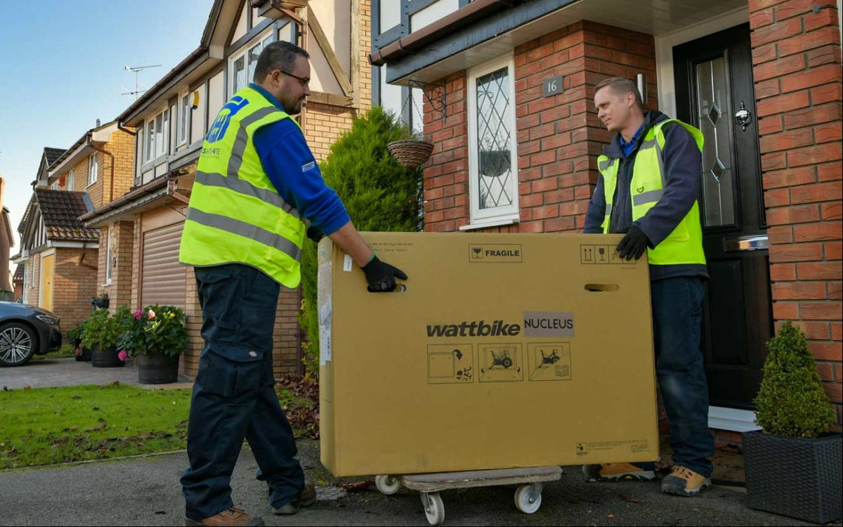 Two workers in high-visibility vests delivering a large box labeled 'Wattbike' in a residential area.